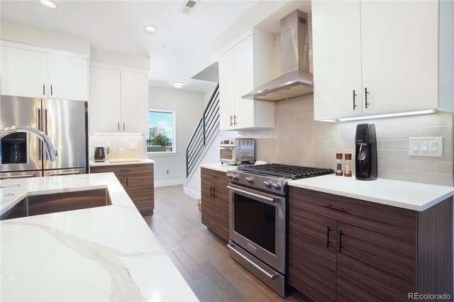 kitchen featuring high end appliances, white cabinetry, backsplash, wood-type flooring, and wall chimney exhaust hood