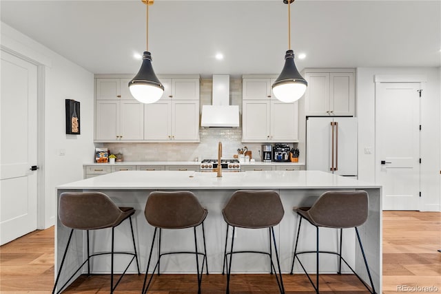 kitchen featuring high quality appliances, premium range hood, an island with sink, decorative light fixtures, and light hardwood / wood-style floors