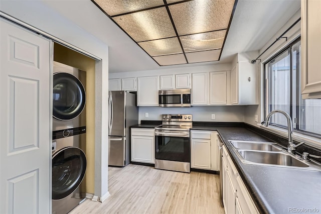kitchen with appliances with stainless steel finishes, a sink, stacked washer / dryer, dark countertops, and light wood-style flooring