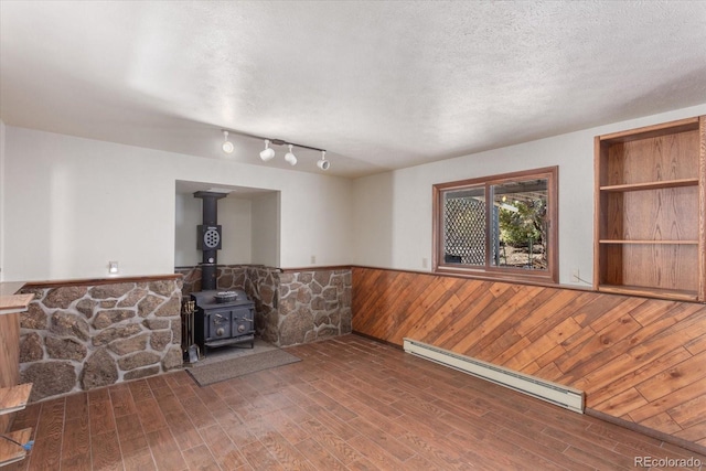 unfurnished living room with a textured ceiling, a baseboard heating unit, wood finished floors, wainscoting, and a wood stove