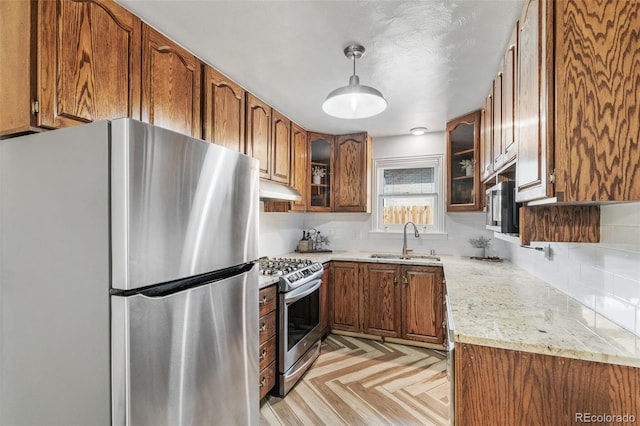 kitchen featuring a sink, under cabinet range hood, appliances with stainless steel finishes, brown cabinetry, and glass insert cabinets