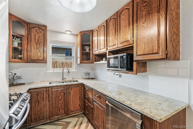 kitchen with a sink, stainless steel appliances, brown cabinetry, glass insert cabinets, and light stone countertops