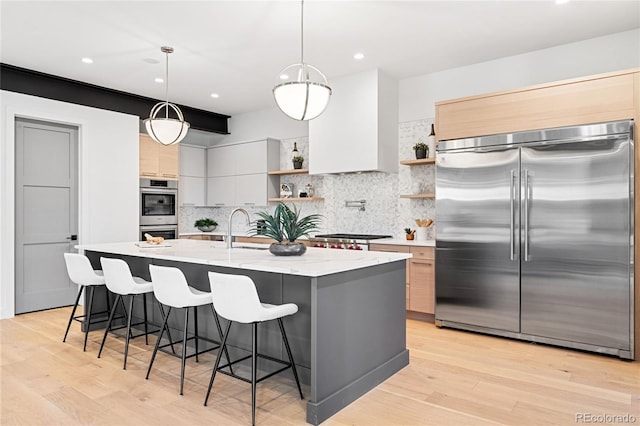 kitchen with light stone countertops, hanging light fixtures, stainless steel appliances, a center island with sink, and light wood-type flooring