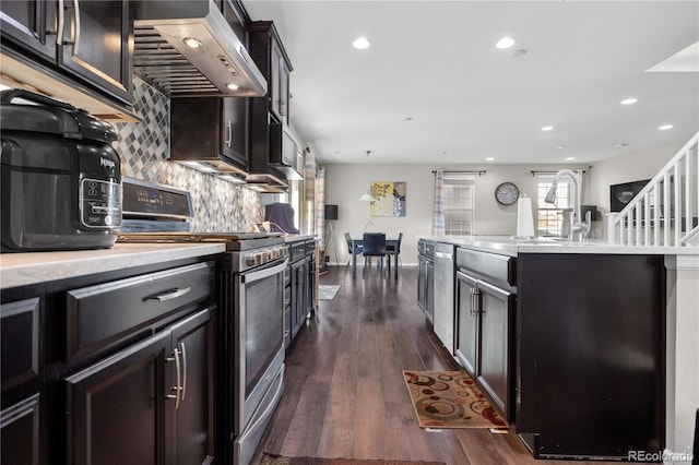 kitchen with wall chimney exhaust hood, dark wood-type flooring, decorative backsplash, a center island with sink, and appliances with stainless steel finishes