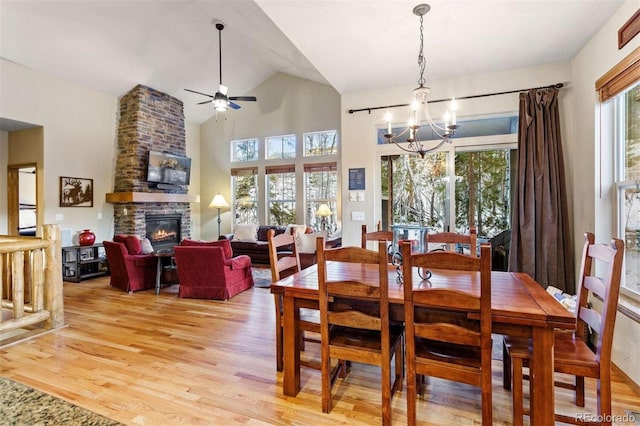 dining space featuring ceiling fan with notable chandelier, high vaulted ceiling, a stone fireplace, and light wood-style flooring