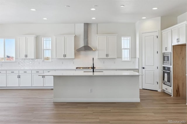 kitchen with stainless steel appliances, white cabinetry, a center island with sink, and wall chimney range hood