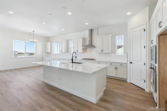 kitchen featuring wall chimney exhaust hood, a kitchen island with sink, sink, and white cabinets