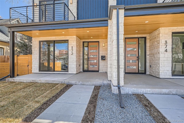 doorway to property with stone siding, a balcony, a porch, and fence