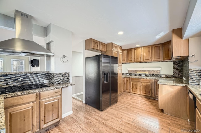 kitchen with light hardwood / wood-style floors, decorative backsplash, black appliances, wall chimney range hood, and dark stone countertops