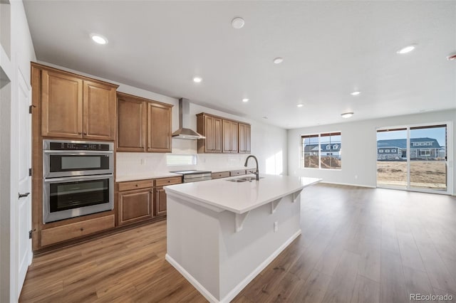 22384 E 49th Pl, Aurora, CO 80019 - kitchen with wall chimney exhaust hood, appliances with stainless steel finishes, light wood-type flooring, brown cabinets, and a center island with sink