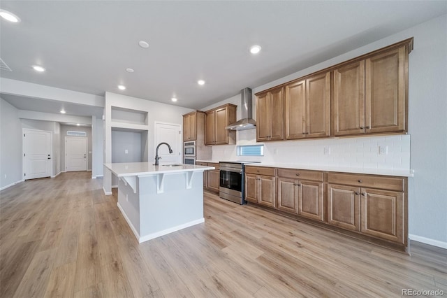 22384 E 49th Pl, Aurora, CO 80019 - kitchen featuring stainless steel appliances, wall chimney range hood, light wood-type flooring, a center island with sink, and brown cabinets