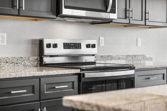 kitchen with a textured wall, appliances with stainless steel finishes, gray cabinets, and light stone counters