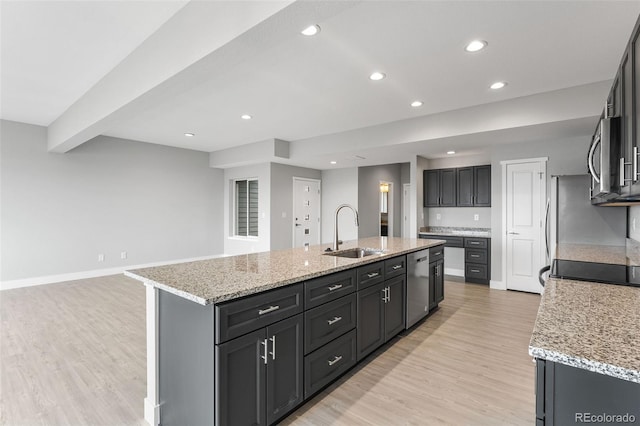 kitchen with light stone countertops, a kitchen island with sink, stainless steel appliances, dark cabinetry, and a sink