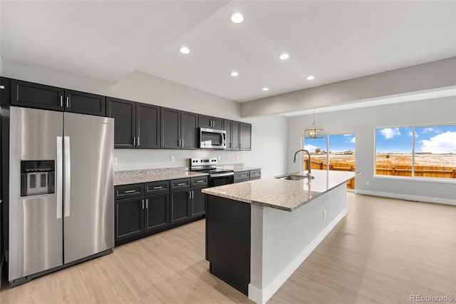 kitchen featuring light stone counters, appliances with stainless steel finishes, a kitchen island with sink, a sink, and dark cabinets