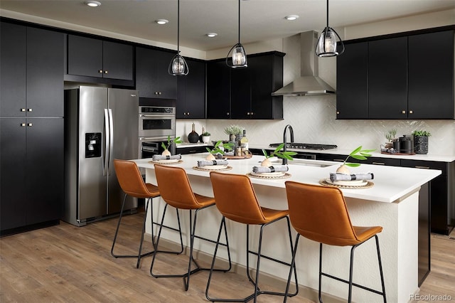 kitchen with a kitchen island with sink, stainless steel appliances, wall chimney exhaust hood, and light hardwood / wood-style floors