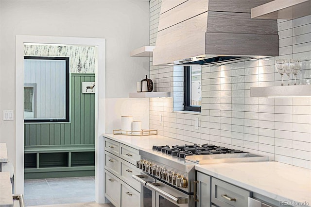 kitchen with range with two ovens, open shelves, decorative backsplash, light stone countertops, and wall chimney exhaust hood