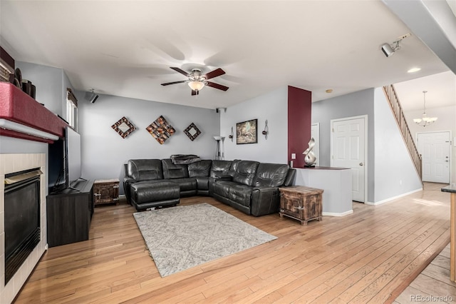 living area featuring a tiled fireplace, ceiling fan with notable chandelier, light wood-style floors, and baseboards