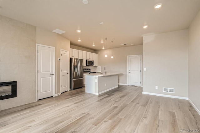 kitchen featuring a center island with sink, a tiled fireplace, stainless steel appliances, pendant lighting, and white cabinetry