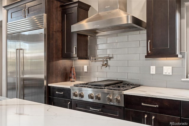 kitchen featuring light stone counters, tasteful backsplash, dark brown cabinets, stainless steel appliances, and wall chimney range hood