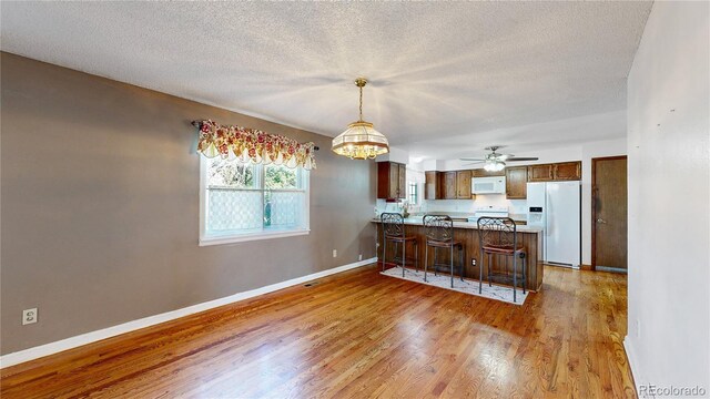 kitchen featuring hardwood / wood-style floors, ceiling fan with notable chandelier, white appliances, kitchen peninsula, and a breakfast bar