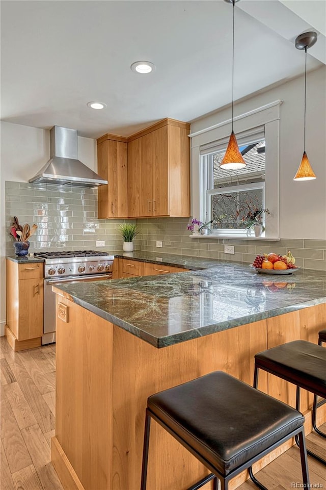 kitchen with a peninsula, stainless steel stove, wall chimney range hood, dark stone counters, and light wood-style flooring