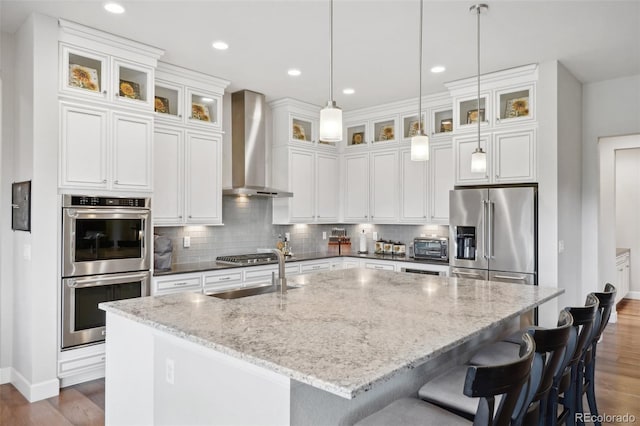 kitchen featuring sink, wall chimney range hood, light stone counters, a kitchen island with sink, and appliances with stainless steel finishes