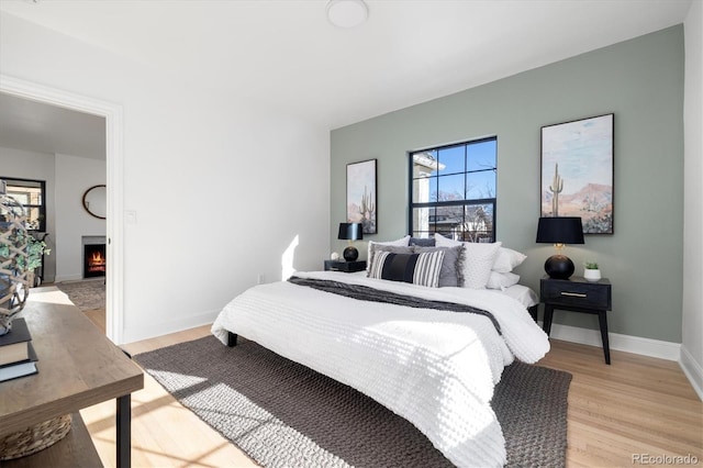 bedroom with light wood-style floors, a warm lit fireplace, and baseboards