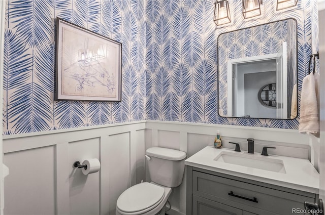 half bathroom with vanity, wainscoting, toilet, and wallpapered walls
