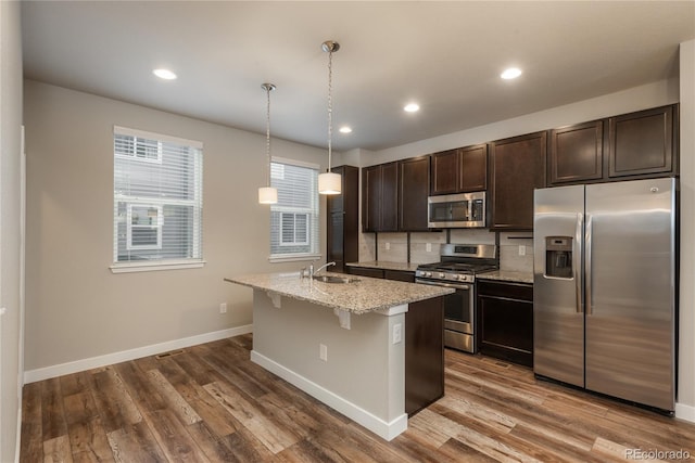 kitchen featuring stainless steel appliances, light stone counters, pendant lighting, a kitchen island with sink, and dark brown cabinets