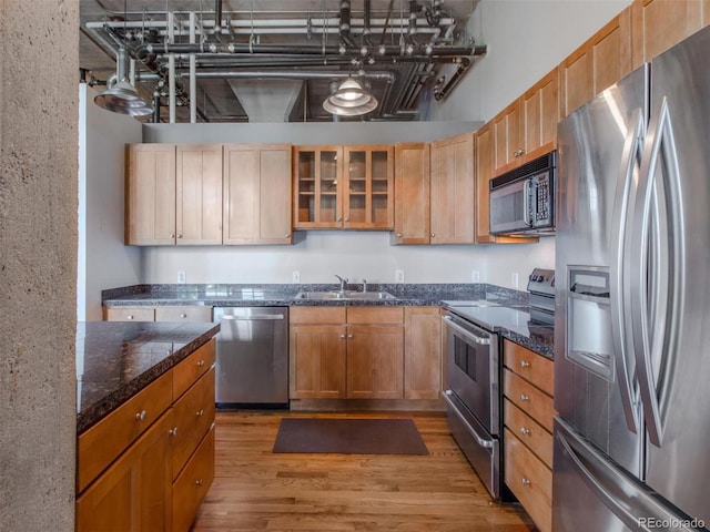 kitchen featuring light wood-style flooring, stainless steel appliances, a sink, dark stone counters, and glass insert cabinets