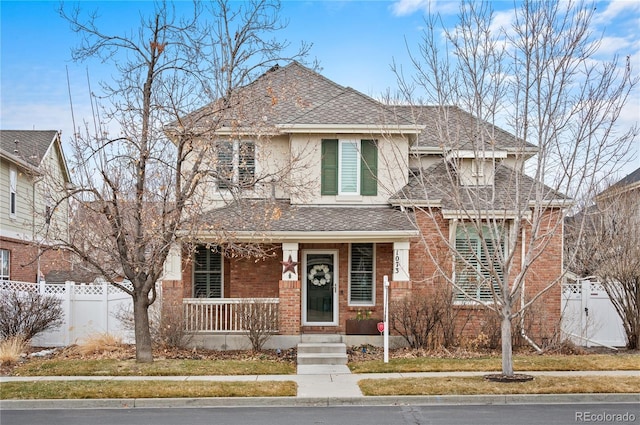 view of front of house with a shingled roof, covered porch, brick siding, and fence