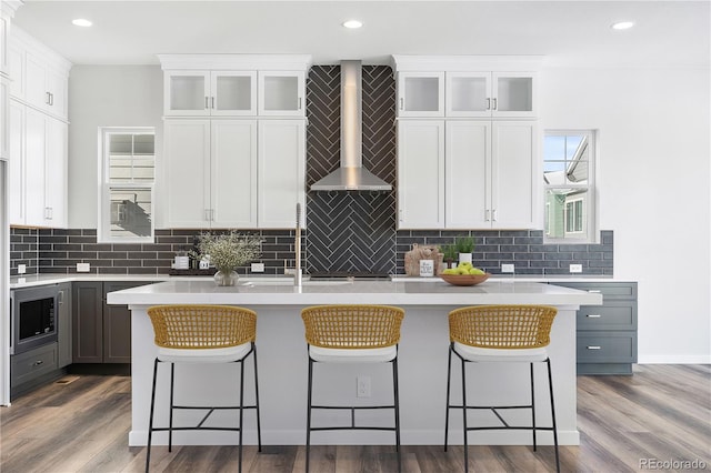 kitchen featuring a kitchen bar, gray cabinetry, a kitchen island with sink, wall chimney range hood, and white cabinets