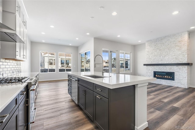 kitchen featuring a stone fireplace, an island with sink, wall chimney exhaust hood, high end stainless steel range, and sink