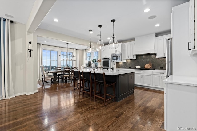 kitchen with a center island with sink, hanging light fixtures, stainless steel appliances, custom range hood, and white cabinets