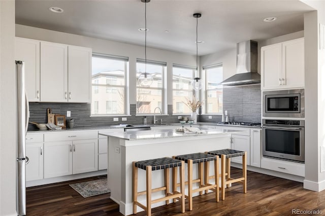 kitchen featuring wall chimney exhaust hood, white cabinetry, light stone counters, a kitchen island, and stainless steel appliances