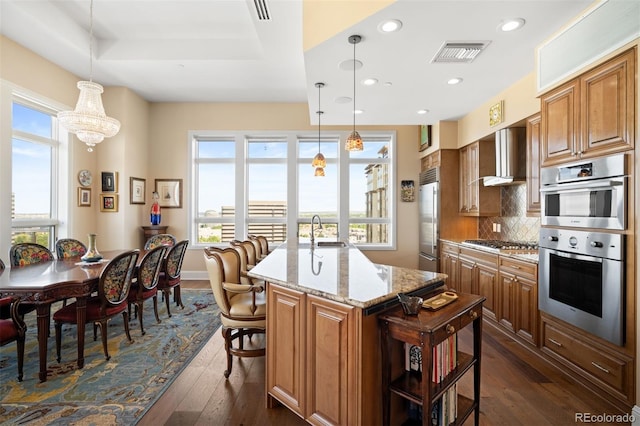 kitchen with visible vents, wall chimney exhaust hood, appliances with stainless steel finishes, hanging light fixtures, and light stone countertops