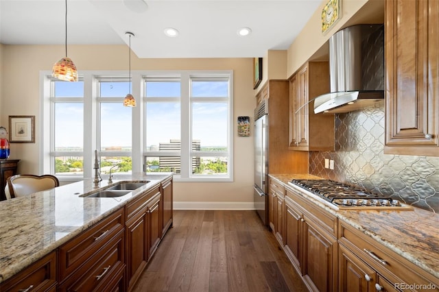 kitchen featuring light stone counters, a sink, appliances with stainless steel finishes, brown cabinets, and wall chimney exhaust hood