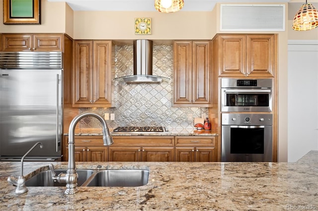 kitchen with light stone counters, stainless steel appliances, hanging light fixtures, a sink, and wall chimney range hood