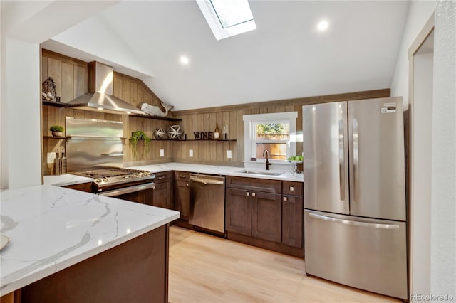 kitchen with vaulted ceiling with skylight, sink, wall chimney exhaust hood, appliances with stainless steel finishes, and light stone countertops