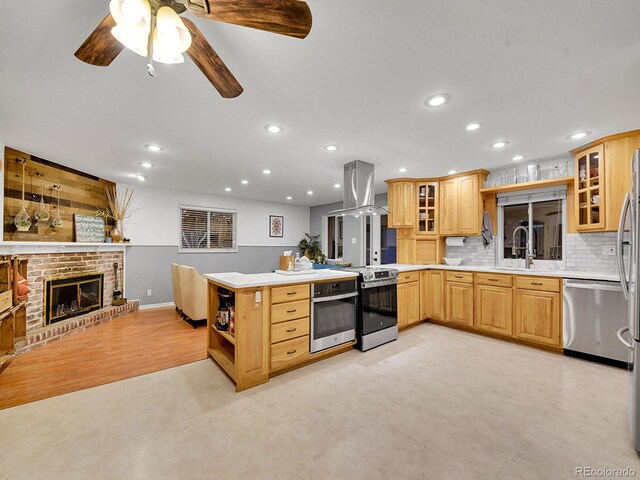 kitchen with backsplash, a brick fireplace, kitchen peninsula, island exhaust hood, and stainless steel appliances