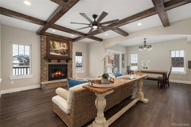 living room with dark wood-type flooring, coffered ceiling, a stone fireplace, beamed ceiling, and ceiling fan with notable chandelier