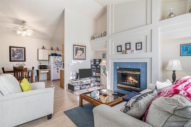 living room featuring high vaulted ceiling, light hardwood / wood-style floors, a tile fireplace, and ceiling fan