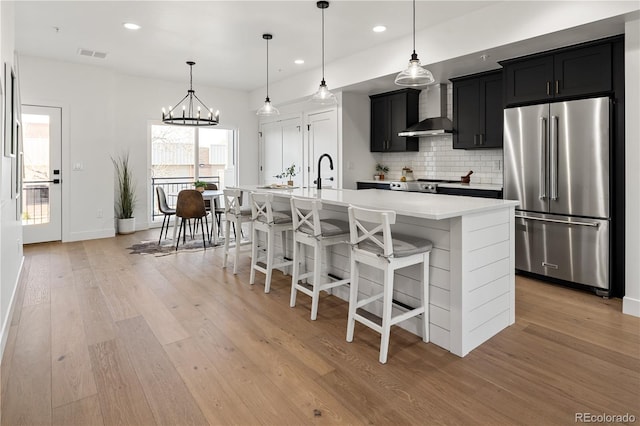 kitchen featuring wall chimney exhaust hood, visible vents, high end refrigerator, light countertops, and dark cabinetry