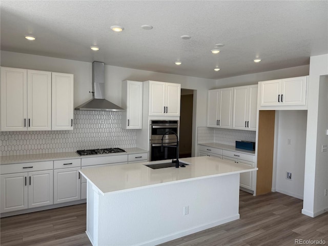 kitchen with wall chimney exhaust hood, gas stovetop, double oven, an island with sink, and white cabinets