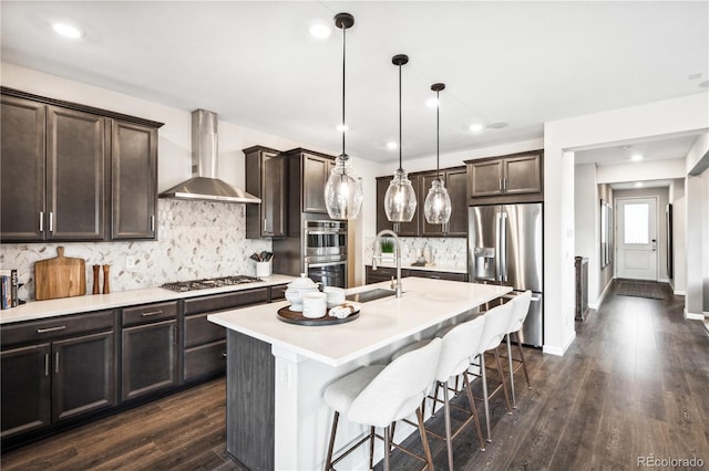 kitchen featuring sink, appliances with stainless steel finishes, a kitchen island with sink, hanging light fixtures, and wall chimney exhaust hood