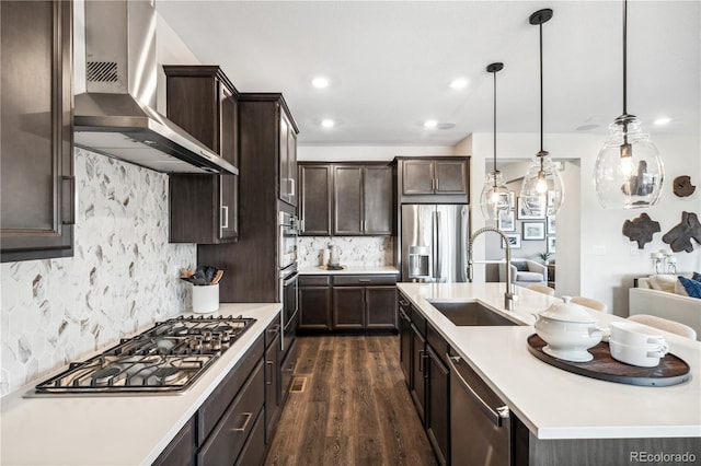 kitchen with sink, a kitchen island with sink, hanging light fixtures, stainless steel appliances, and wall chimney exhaust hood