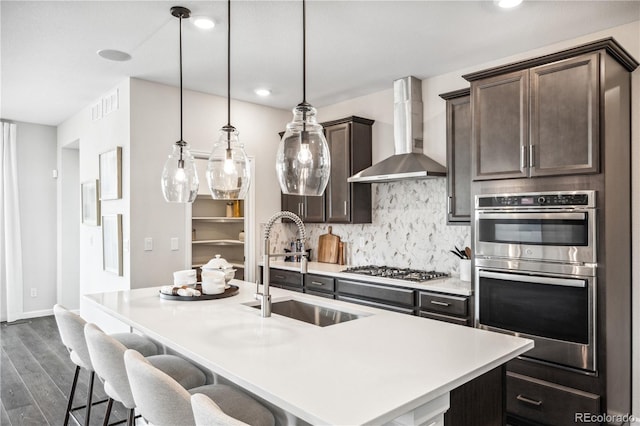 kitchen featuring appliances with stainless steel finishes, wall chimney exhaust hood, decorative light fixtures, and a center island with sink