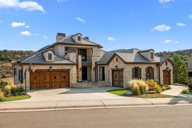 french country style house featuring a balcony, stone siding, a chimney, concrete driveway, and an attached garage