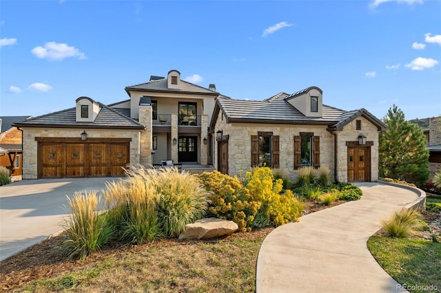 french country inspired facade featuring stone siding, a garage, stucco siding, concrete driveway, and a balcony