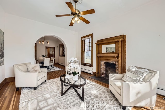 living area featuring arched walkways, wood finished floors, a baseboard radiator, ceiling fan, and a fireplace